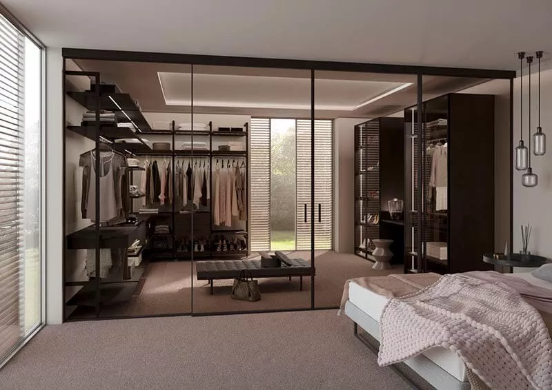 Auf diesem Bild wird ein Ankleidezimmer in Brauntönen durch einen Raumteiler vom eigentlichen Schlafzimmer abgegrenzt. Die Raumteiler sind durchsichtig und geschlossen. Man kann jedoch alles dahinter durch leicht getönte Schieben sehen. Im Ankleidezimmer befinden sich einige Regale in einem Säulensystem und weitere Einbauschränke für besonders wertvolle Dinge. In der Mitte ist eine kleine Ledercouch.