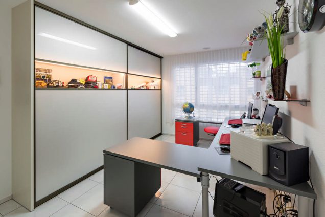 Ein Einbauschrank, mit geschlossenen sehr breiten Schiebetüren in einem Büroraum. Das Abiente wirkt wie in einem Homeoffice. Die Hauptfarbe auf dem Bild ist ein edles Dunkelgrau. Die Schiebetüren des Einbauschrankes bestehen oben und unten aus hellgrauen Elementen, eingefasst in dunklem Holz. In der Mitte der Schiebetür befindet sich ein helles beleuchtetes Band aus Glas, hinter dem sich mehrere Ausstellungsstücke befinden. Das Büro ist teilweise mit roten Elementen ergänzt.