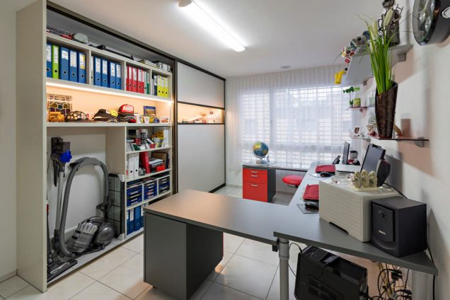 Hier sehen wir einen zu weiten Teilen geöffneten Einbauschrank im Homeoffice. In dem Schrank befindet sich ein Staubsauger und sehr viele bunte Ordner. In der mitte ist ein beleuchtetes Fach mit sehr ansehnlichen Ausstellungsstücken. Dieses beleuchtete Fach ist hinter einem durchsichtigen Glas, wenn die Schiebetüren des Einbauschrankes geschlossen sind. Weiters befindet sich auf dem Foto noch ein Tisch und ein paar kleine Kästen, welche zum HomeOffice gehören.