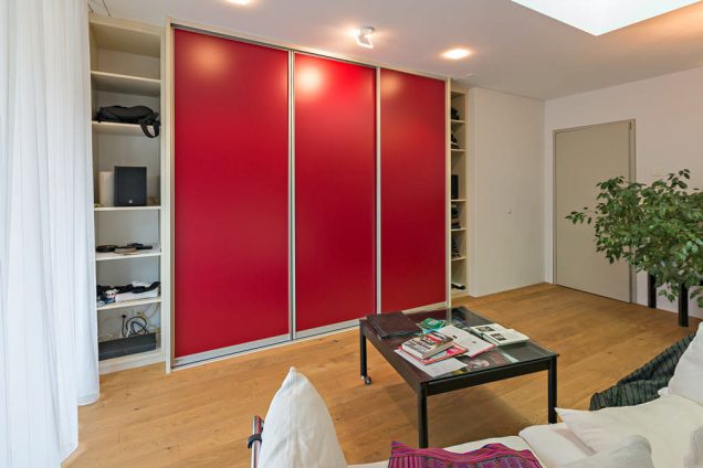 Dieser Einbauschrank besteht aus 5 Teilen, die sich aneinander reihen Die äußersten 2 sind etwas nach hinten versetzt und haben keine Türe. Die 3 in der Mitte haben rote Schiebetüren, welche von den Spots an der Decke angestrahlt werden. In den Regalen rechts und links befinden sich diverse Alltagsgegenstände aus dem Wohnzimmer. In der Mitte ist eine der Schiebetüren geöffnet und dahinter sind mehrere Schubladen und Regale zu sehen, die mit diversen Kleidungsstücken gefüllt sind.
