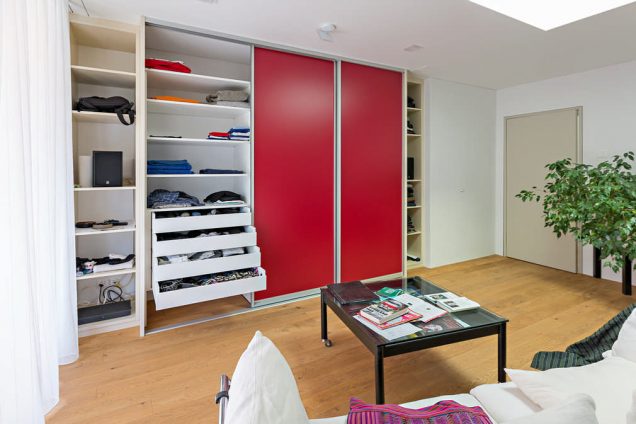 Dieser Einbauschrank besteht aus 5 Teilen, die sich aneinander reihen Die äußersten 2 sind etwas nach hinten versetzt und haben keine Türe. Die 3 in der Mitte haben rote Schiebetüren, welche von den Spots an der Decke angestrahlt werden. In den Regalen rechts und links befinden sich diverse Alltagsgegenstände aus dem Wohnzimmer. In der Mitte ist eine der Schiebetüren geöffnet und dahinter sind mehrere Schubladen und Regale zu sehen, die mit diversen Kleidungsstücken gefüllt sind.