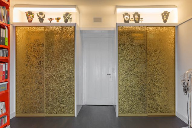 Der hier dargestellte Einbauschrank besteht aus geschlossenen goldenen Schiebetüren, welche richtig schimmern. Über den Schiebetüren befinden sich zwei Glasvitrinen, welche von innen beleuchtet sind und darin befinden sich mehrere Büsten mit den unterschiedlichsten Ketten. In der Mitte ist eine weiße Tür, die augenscheinlich in einen anderen Raum führt.