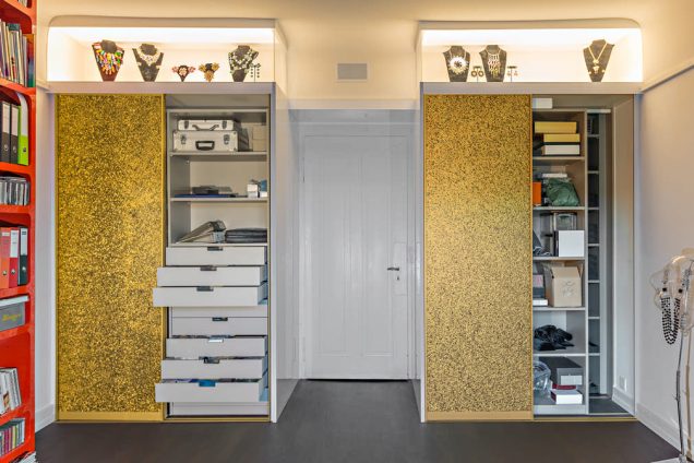 Diese Sonderanfertigung eines Einbauschranke besteht aus goldenen Schiebetüren. Darüber befindet sich eine von hinten beleuchtete Glasvitrine in der verschiedene Büsten mit Schmuck ausgestellt sind. Im Zentrum des Einbauschrankes ist ein raumhoher Durchgang, welcher an einer weißen Türe endet. Die linke Schrankhälfte ist teilweise geöffnet und darin befinden sich viele Schubladen und Regale für Taschen und Schmuck. Auch die rechte Seite ist teilweise geöffnet. Darin befindet sich ein fast raumhohes Regal gefüllt, in dem alle möglichen Alltagsgegenstände einsortiert worden sind.