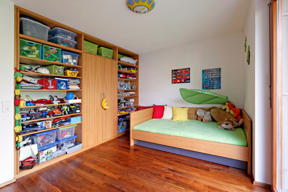 Dieser Einbauschrank besteht hauptsächlich aus offenen Regalen, welche die gesamte Wand ohne Luftraum einnehmen. Die Regale sind gut gefüllt mit allerlei Spielsachen und Boxen. In der Mitte befindet sich ein Drehtürenschrank mit einem gelben Mondanhänger am Griff. Rechts ist ein Kinderbett mit grünem Bettbezug und bunten Kissen.