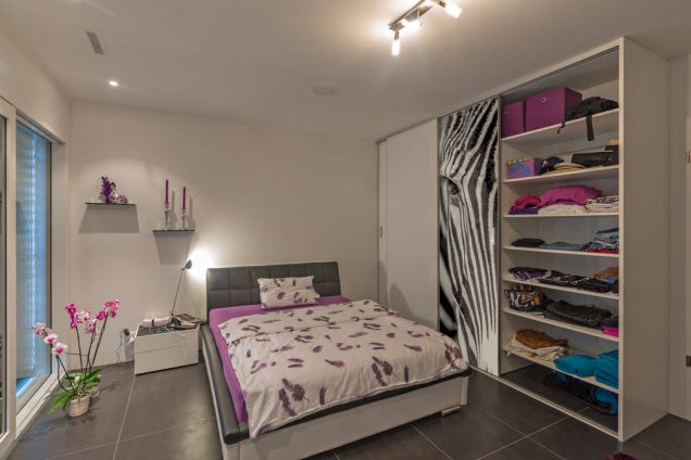 Dargestellt ist ein Schlafzimmer mit einem Bett als Sonderanfertigung in violett- und Anthrazittönen. Rechts davon ist ein Einbauschrank mit einer weißen Schiebetür und einer, auf der ein Zebrakopf abgebildet ist. Das ganze Ambiente wirkt sehr modern und aufregend. In dem Einbauschrank ist ein Regal abgebildet, welches mit allen möglichen Alltagsgegenständen gefüllt ist.
