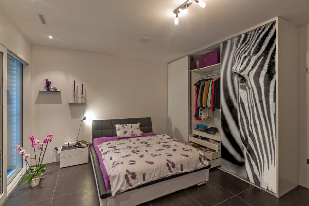 Dargestellt ist ein Schlafzimmer mit einem Bett als Sonderanfertigung in violett- und Anthrazittönen. Rechts davon ist ein Einbauschrank mit einer weißen Schiebetür und einer, auf der ein Zebrakopf abgebildet ist. Das ganze Ambiente wirkt sehr modern und aufregend. In dem Einbauschrank befinden sich mehrere Schubladen und Kleiderstangen gefüllt mit allen möglichen Kleidungsstücken. Im oberen Bereich ist noch ein zusätzliches Regal, das noch mehr Stauraum bietet.